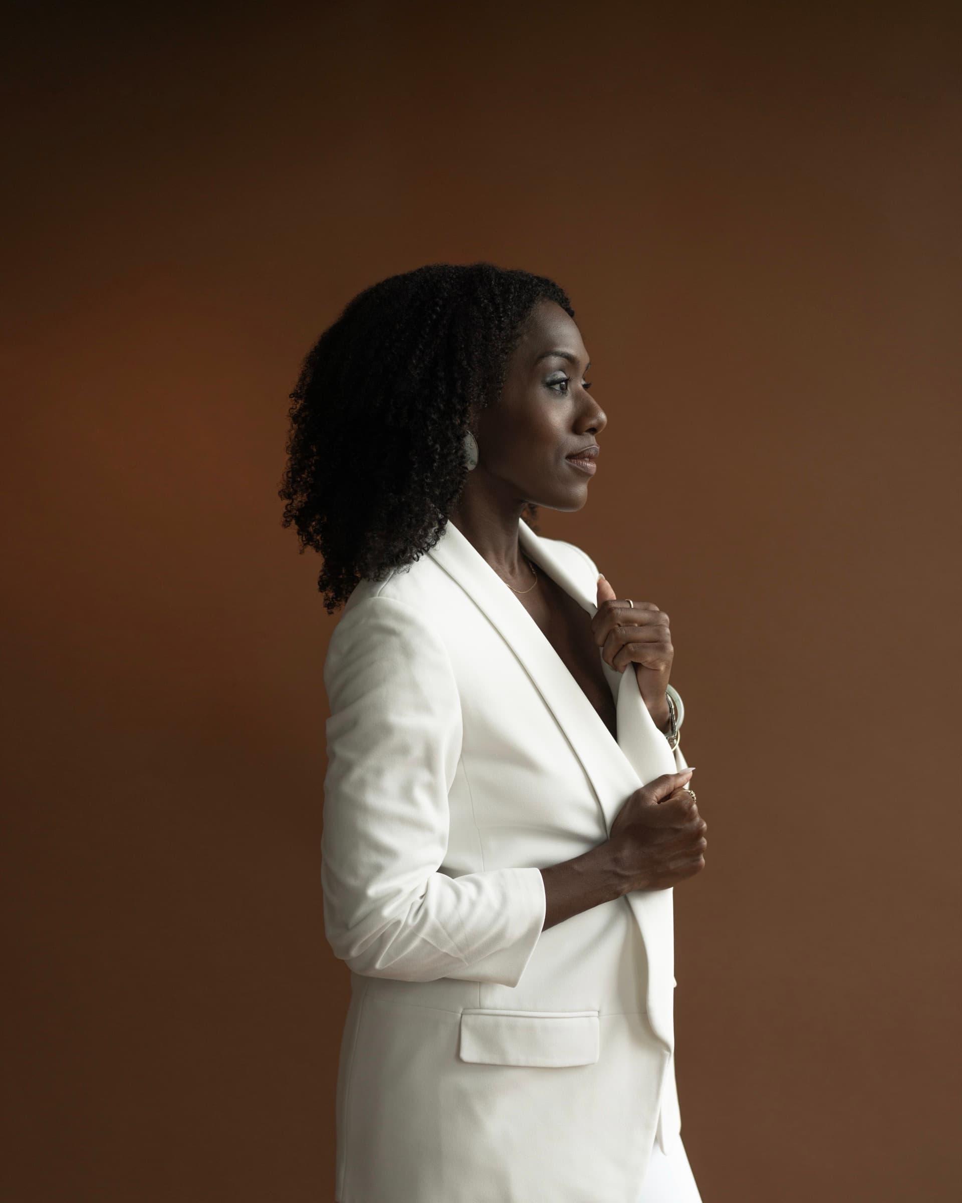 Alicia Waller in profile wearing a cream suit against a deep brown backdrop.